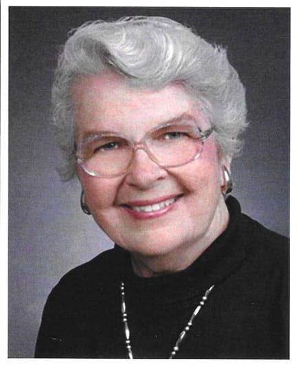 Picture of older lady with glasses and gray hair, wearing a black mock neck sweater and a gold necklace.
